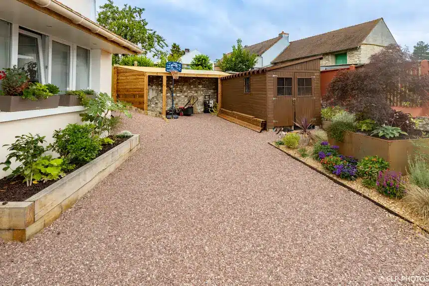 Allée gravillonnée bordée de jardinières en bois menant aux abris de jardin en bois